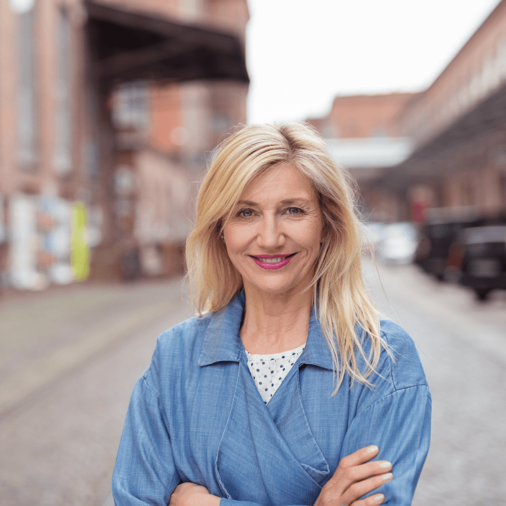Confident woman with soft blonde hair and natural highlights, smiling warmly while standing on a cobbled street in a relaxed blue outfit. A beautiful example of expert cutting and highlighting, reflecting the personalised styling services at Baileys Hairdressing in Grantham.