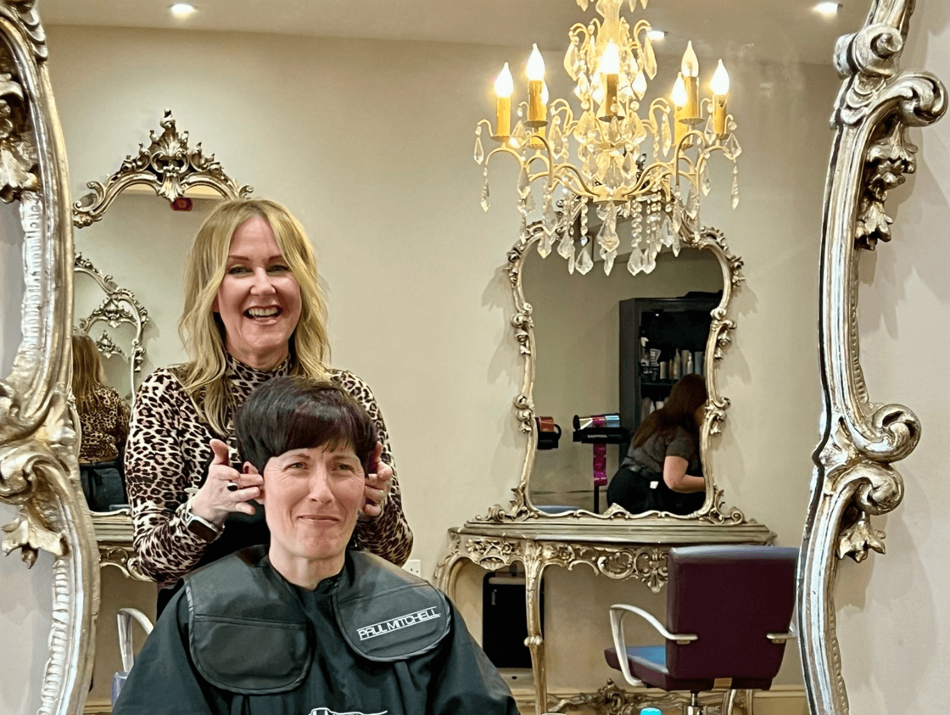 One of the friendly stylists at Baileys Hairdressing smiling with a client in the salon, showcasing the warm and elegant atmosphere with ornate mirrors and chandeliers.