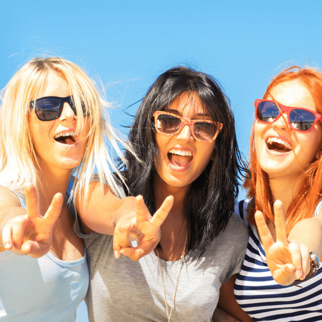 Three vibrant women with blonde, black, and copper-red hair tones, smiling and making peace signs under a bright blue sky. A fun, feel-good image celebrating bold, beautiful hair colour results, perfect for showcasing the creativity and vibrancy of professional colour services at Baileys Hairdressing in Grantham.